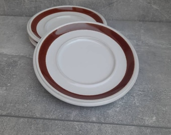 May include: A stack of white ceramic plates with a wide, brown band around the rim. The plates are stacked on a gray surface. The plates are a simple, classic design, suitable for everyday use.