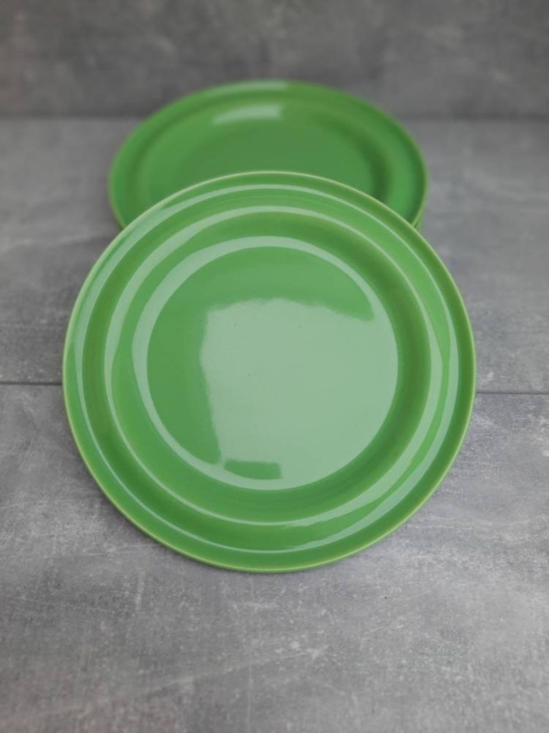 May include: Two bright green ceramic plates. The plates are round with a slightly raised rim and concentric rings. The plates are a solid, glossy green color. The plates are on a gray surface.