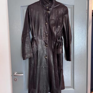 Op de afbeelding: Een donkerbruine leren trenchcoat met een hoge kraag, lange mouwen en een knoopsluiting aan de voorzijde. De jas is voorzien van een bijpassende leren ceintuur in de taille en verticale naaddetails over het hele kledingstuk voor een pasvorm.