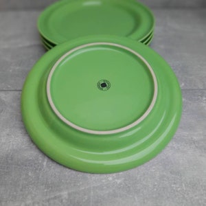 May include: A stack of four bright green ceramic plates. The plates have a rounded shape with a slightly raised rim. The underside of one plate is visible, showing a white rim and a small, circular logo.