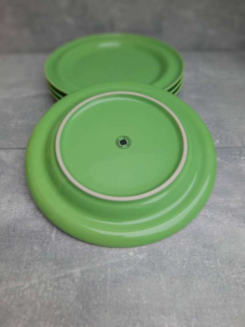 May include: A stack of four bright green ceramic plates. The plates have a rounded shape with a slightly raised rim. The underside of one plate is visible, showing a white rim and a small, circular logo.