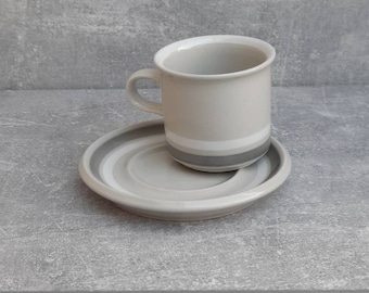 May include: A ceramic coffee cup and saucer set. The cup is light gray with a white rim and a dark gray band. The saucer has concentric gray rings. The set is designed for serving hot beverages.