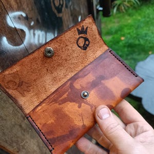 Könnte beinhalten: Eine braune Ledergeldbörse mit einem Kronen- und Totenkopf-Logo. Die Geldbörse hat einen Used-Look und wird mit einem Druckknopf geschlossen.