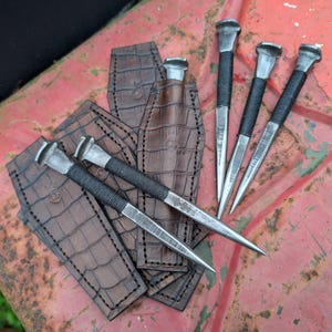 May include: Handmade metal awls with black wrapped handles and brown leather sheaths. The awls have a pointed tip and a flat head. The leather sheaths have a crocodile pattern and are stitched around the edges. The awls and sheaths are displayed on a red surface.