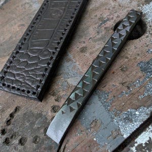 May include: A dark brown leather sheath with a textured crocodile-skin pattern and visible stitching alongside a long, narrow metal tool featuring a series of triangular indentations along its top edge and a flat, beveled blade at the tip.