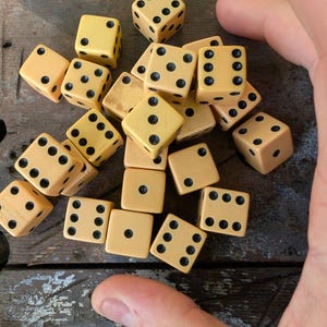Può includere: Una collezione di dadi a sei facce color crema con punti neri sparsi in un mucchio. Ogni dado presenta angoli arrotondati e marcature numeriche standard da uno a sei, mostrando varie orientazioni e motivi di punti nel gruppo.