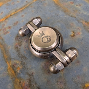 May include: A metallic fidget spinner with a weathered bronze finish, featuring a central circular cap engraved with a skull wearing a crown. The device has two arms extending from the center, each tipped with a hexagonal nut and a rounded end cap.