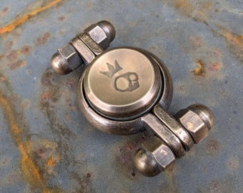 May include: A metallic fidget spinner with a weathered bronze finish, featuring a central circular cap engraved with a skull wearing a crown. The device has two arms extending from the center, each tipped with a hexagonal nut and a rounded end cap.