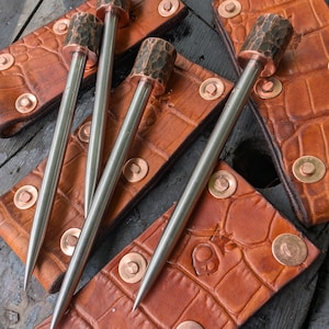 May include: A collection of metal skewers with hammered copper tops, resting on textured brown leather holders with copper rivets. The skewers have a sharp point and a polished metal shaft. The leather holders have a crocodile skin pattern.
