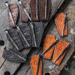 May include: A collection of coffin-shaped leather sheaths in brown, black, and tan, each featuring a crocodile-embossed texture, stitched edges, and a stamped crown logo, paired with a long, pointed metal nail resting on each individual sheath.