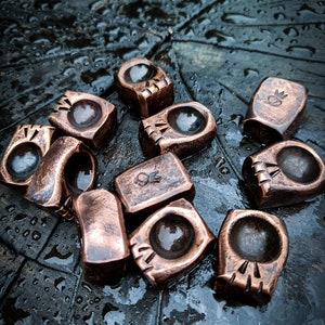 May include: A collection of ten small, copper-coloured metal rings with various designs. The rings have a slightly rounded, rectangular shape and feature different patterns, including a crown, a skull, and a series of lines.