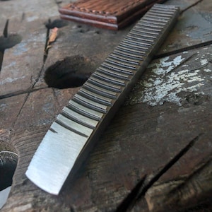 May include: A rectangular metal tool featuring a series of parallel, raised ridges along its length and a smooth, flat, tapered end. The tool has a dark, weathered finish and is positioned diagonally across the frame, showcasing its textured surface.
