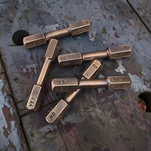 May include: Several brass decision makers are scattered on a weathered wooden surface. Each has a hexagonal head with "YES" and "NO" engraved on opposite sides. The cylindrical body has a series of small holes. The overall color is a warm, aged brass.
