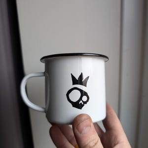 Può includere: Tazza in smalto bianco con bordo nero e un grafico di teschio nero con corona.