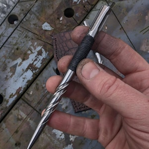 Puede incluir: Un bolígrafo de metal hecho a mano con un diseño en espiral en la parte inferior del cuerpo y un agarre envuelto en negro. El bolígrafo se sostiene en una mano, mostrando su acabado pulido y su punta. El fondo es una superficie de madera desgastada.