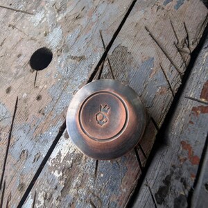 May include: A circular copper-toned metal knob with a brushed finish and a recessed center. The center features a small, stamped icon of a skull wearing a crown. The object has a smooth, rounded edge and a distinct metallic sheen throughout its surface.