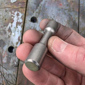 May include: A cylindrical metal tool with a rounded top and bottom, held in a hand. The tool has a narrow, ridged center section and a matte silver finish. The background is a weathered wooden surface.