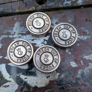 May include: Four silver-colored metal medallions with the text "Fresh Out Of Fucks" and a skull and crown design.