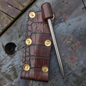 Puede incluir: Una herramienta de cuero marrón con un patrón de cocodrilo, asegurada con remaches dorados. Un mango de madera está unido a una punta de metal. La herramienta está diseñada para trabajos en cuero o artesanías similares.