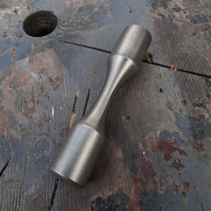 Könnte beinhalten: Ein silberfarbenes Metallwerkzeug mit strukturierter Oberfläche. Das Werkzeug hat eine zylindrische Form mit einer schmalen, sanduhrartigen Mitte. Es liegt auf einer verwitterten Holzoberfläche.