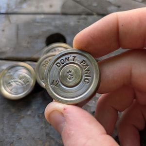 May include: A circular brass-toned metal token featuring the embossed text DON'T PANIC along the upper rim, the number 42 at the bottom, and a central symbol. The token has a polished finish with concentric circular grooves on its surface.