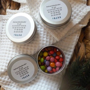 Könnte beinhalten: Adventskalender-Dosen mit Deckeln, jeweils mit den Nummern 1-24 beschriftet. Eine Dose ist geöffnet und zeigt bunte Süßigkeiten. Die Dosen befinden sich auf einem weißen Tuch und einem geflochtenen Korb. Der Text auf dem Etikett lautet "Adventskalender to go".