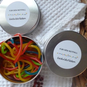 Könnte beinhalten: Drei kleine, silberne Dosen mit weißen Etiketten und farbigem Text. Eine Dose ist geöffnet und zeigt bunte Gummibonbons. Der Text auf den Etiketten lautet "FÜR DEN NOTFALL Geduldsfäden."