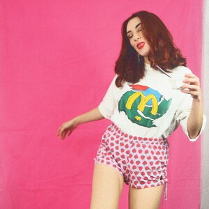 May include: A woman wearing a white t-shirt with a colorful graphic of a stylized "M" and a strawberry print shorts. She is holding a glass with a straw in her hand.