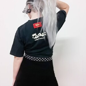 May include: Black t-shirt with a white dragon graphic and the text "Pink Dragon" on the back. The shirt has a red square label with the text "2AF" on it. The person is wearing a black cap and a black mini skirt with a silver studded belt.