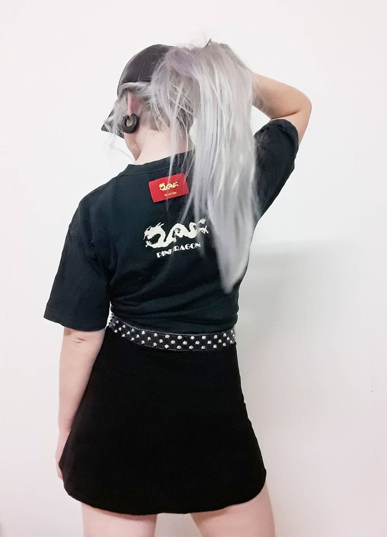 May include: Black t-shirt with a white dragon graphic and the text "Pink Dragon" on the back. The shirt has a red square label with the text "2AF" on it. The person is wearing a black cap and a black mini skirt with a silver studded belt.