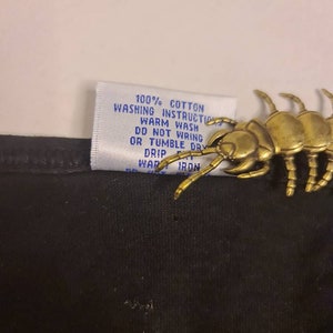 May include: A white tag with blue text attached to a black fabric garment. The tag reads "100% Cotton Washing Instructions Warm Wash Do Not Wring Or Tumble Dry Drip Warm Iron".