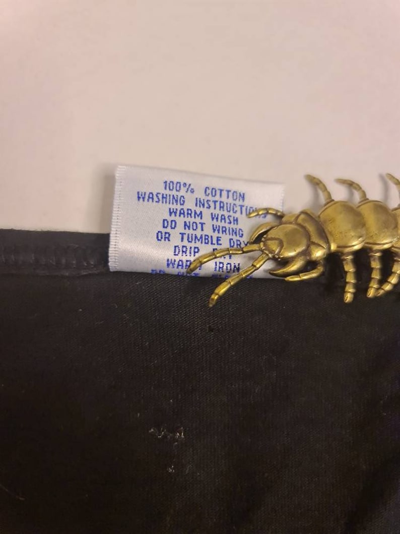 May include: A white tag with blue text attached to a black fabric garment. The tag reads "100% Cotton Washing Instructions Warm Wash Do Not Wring Or Tumble Dry Drip Warm Iron".