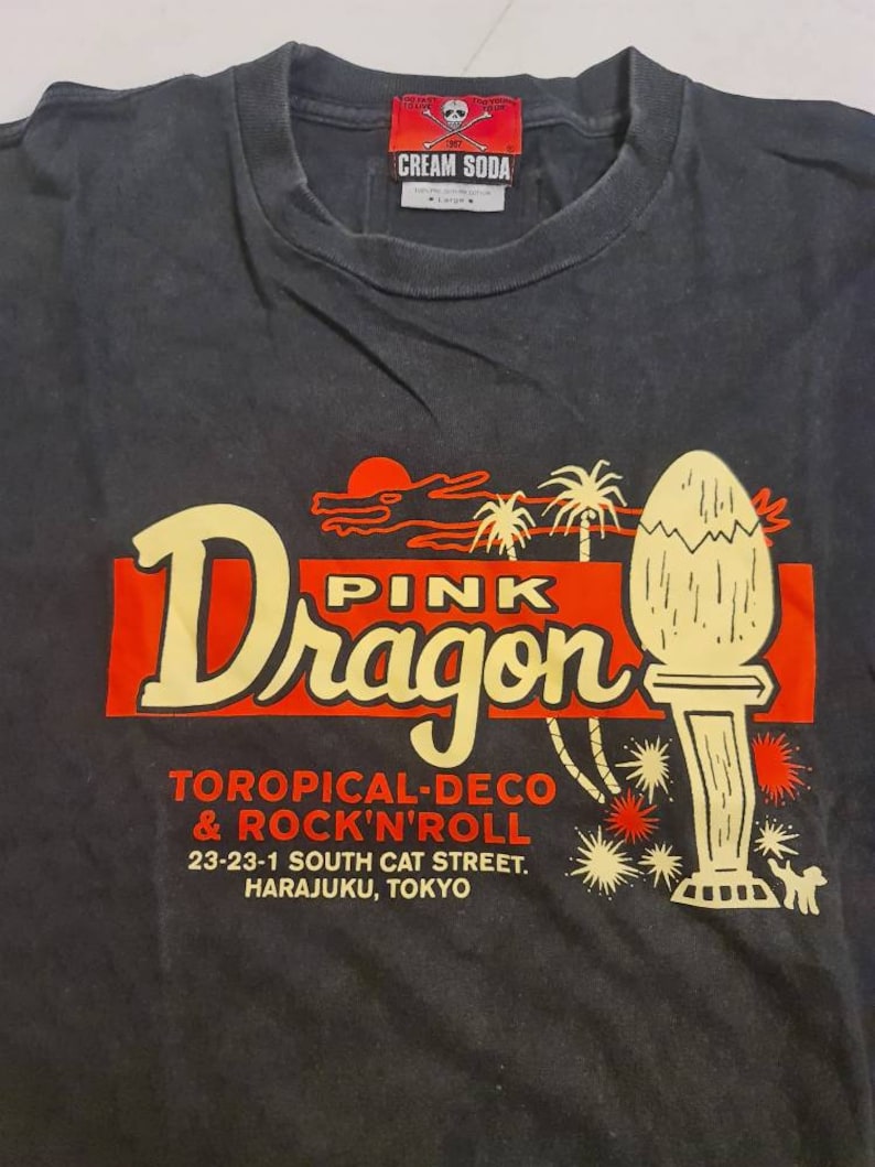May include: Black t-shirt with a red and yellow graphic of a pink dragon with the text "Pink Dragon", "Toropical-Deco & Rock'n'Roll", "23-23-1 South Cat Street. Harajuku, Tokyo".