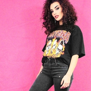 May include: A woman with long, curly, dark red hair is wearing a black t-shirt with a band logo and black jeans. The background is pink.