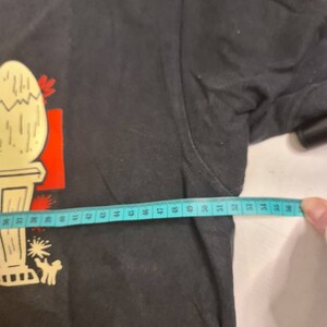 May include: Black t-shirt with a cartoon illustration of a cracked egg on a pedestal. The shirt has a white measuring tape across the bottom with numbers 36 to 56.