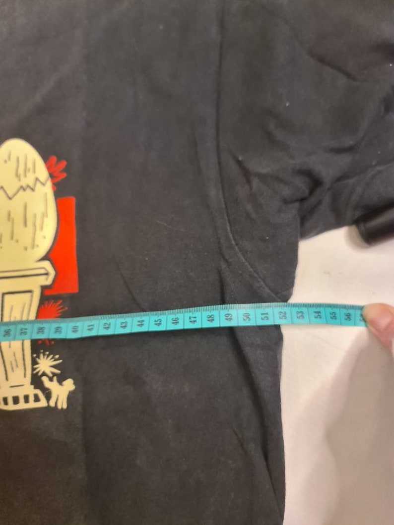 May include: Black t-shirt with a cartoon illustration of a cracked egg on a pedestal. The shirt has a white measuring tape across the bottom with numbers 36 to 56.