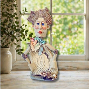 Op de afbeelding: Een grillige keramische sculptuur van een persoon met blauwe ogen, rode lippen en stekelig bruin haar. De figuur draagt een sjaal met patroon en heeft de handen in de zij. De sculptuur is gebroken wit met vleugjes kleur en staat op een houten oppervlak.