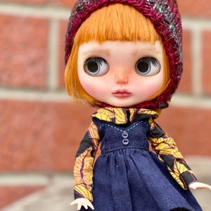 Può includere: Una bambola con i capelli arancioni che indossa un cappello di lana bordeaux con un pon pon giallo, una camicia a fantasia gialla e nera e un vestito blu scuro.