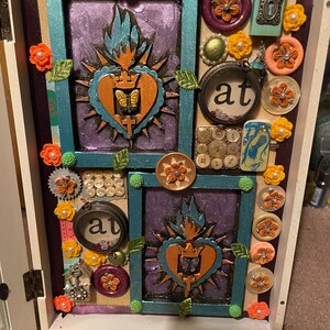 May include: A white wooden box with a teal and purple background. The box is decorated with various embellishments, including buttons, flowers, and a small metal key. The box has two framed images of a heart with a crown and a flame. The text "at" is visible on the box.
