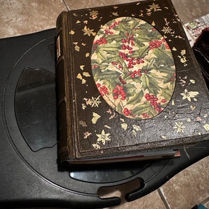 May include: A brown leather-bound book with a gold-leaf design and a circular image of holly leaves and red berries.