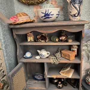 May include: A small, blue, wooden cabinet with shelves and a door. The cabinet is filled with miniature items, including a teapot, a bowl, and a jar. The cabinet is sitting on a table with a floral wallpaper background.
