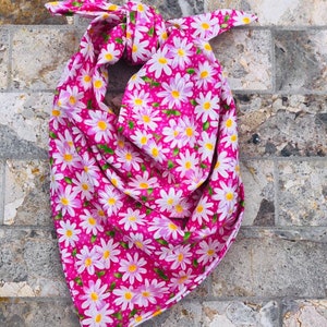 May include: A pink bandana with a white daisy print. The bandana is tied in a knot and is lying on a stone surface.