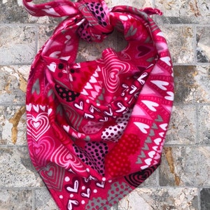 Peut inclure: Bandana rose avec un motif de cœurs. La bandana est nouée et présente une variété de cœurs roses, blancs et gris.