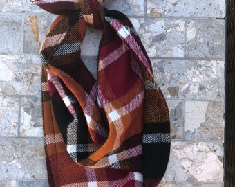 May include: A plaid bandana with a brown, black, white, and burgundy color scheme. The bandana is tied in a knot and is hanging on a stone wall.