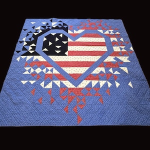 May include: A blue, red, and white quilt featuring a heart-shaped American flag design. The flag is composed of red and white stripes and a blue field with white stars. The quilt has a geometric pattern and is displayed on a black background.