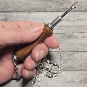 May include: A seam ripper with a wooden handle and a silver metal blade. The blade has a red ball at the tip. A silver chain is attached to the handle. The tool is designed for removing stitches from fabric.