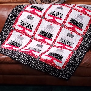 May include: A handmade quilt featuring a repeating pattern of cakes on red stands against a white background. The cakes have black and white patterns. The quilt has a black border with white polka dots. The quilt is draped over a brown leather couch.