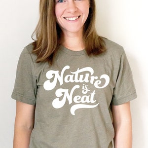 May include: A woman wearing a green t-shirt with the text "Nature is Neat" in white lettering. The shirt has a relaxed fit and short sleeves.
