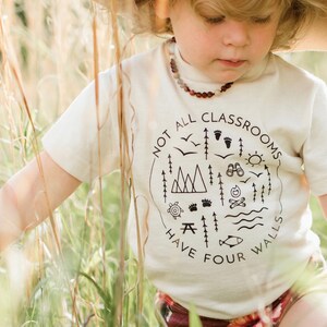 Puede incluir: Un ni&ntilde;o con una camiseta blanca con un gr&aacute;fico negro que dice "No todas las aulas tienen cuatro paredes" y un sombrero marr&oacute;n est&aacute; de pie en un campo de hierba alta.