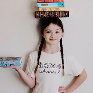 May include: A young girl with long brown braids is wearing a white t-shirt that says "home schooled". She is holding a blue book with colorful flowers on the cover and has a stack of books balanced on her head. The books on her head are a mix of colors and have titles like "Moby Dick" and "Classic Poetry".